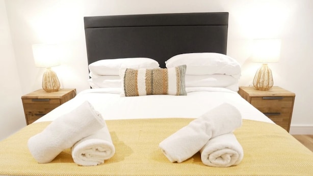 Un lit bien fait, avec du linge de lit blanc et deux serviettes enroulées posées sur une couverture jaune. Il y a deux tables de chevet avec des lampes, une de chaque côté du lit.