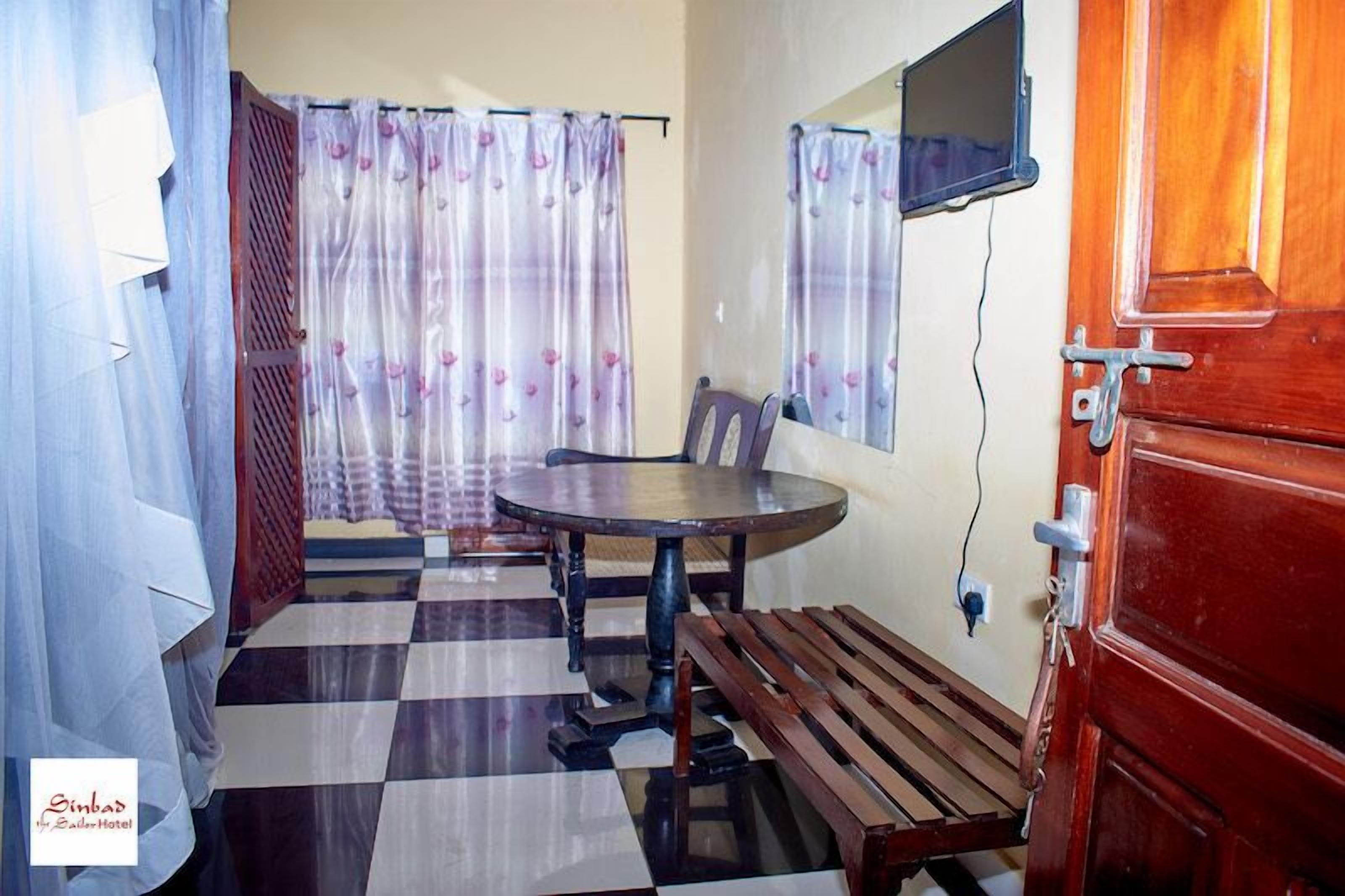 A room with a checkered floor, a round table with chairs, a wooden bench, a wooden door, and a window with a curtain.