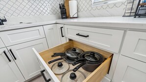 Una cucina con mobili bianchi, un cassetto in legno contenente pentole e padelle, un porta carta da cucina e un portacoltelli.