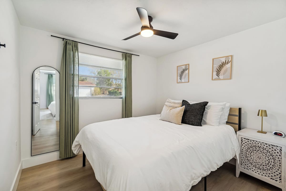 Un dormitorio con cama, ventilador de techo, mesita de noche con lámpara y un cuadro en la pared.
