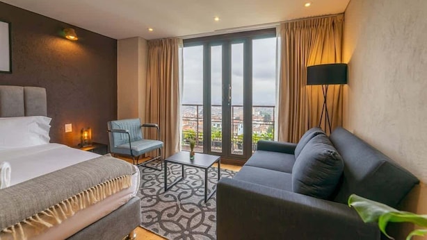 Een hotelkamer met een bed, een slaapbank, een stoel, een klein tafeltje en een groot raam met gordijnen.