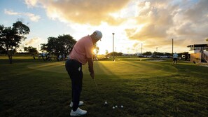 Ein Golfplatz mit einer Person, die zum Putt ansetzt, Bäumen und einem Sonnenuntergang im Hintergrund.
