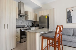 A modern kitchen with wooden cabinets, a stainless steel refrigerator, a gas stove, and a bar area with wooden stools. - NOK Modern 1BR Apartment with Terrace in Vistacana (Punta Cana)
