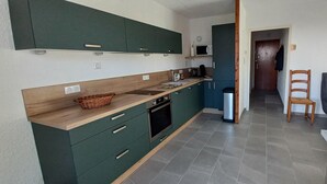 A compact kitchen with green cabinets, a microwave, and a trash bin. There is a wooden table and two wooden chairs in the adjacent living area. - "Aux Aurores" – Charming Apartment with Mountain View and Balcony (Embrun)