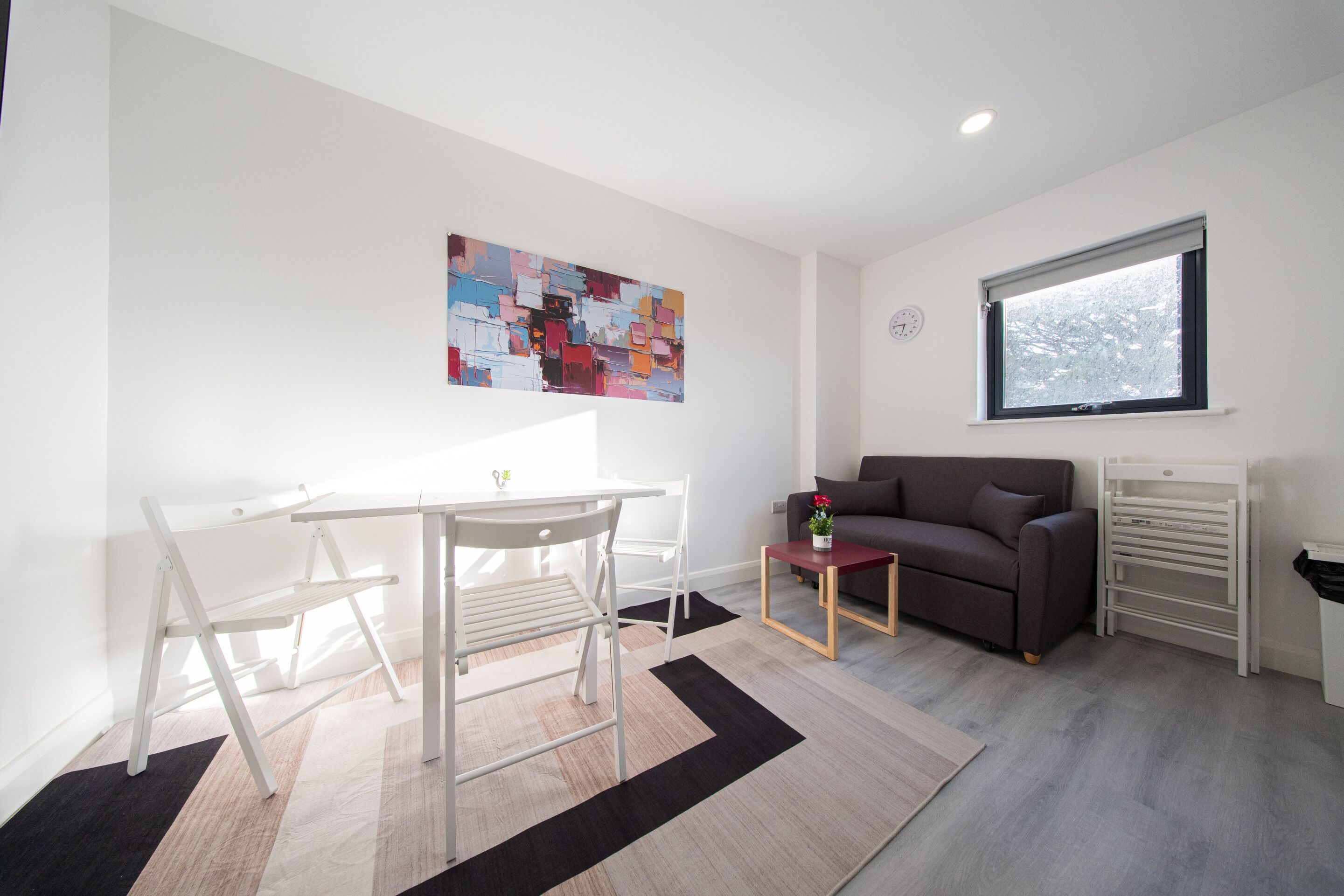 A modern living space with a white table, chairs, a grey sofa, a painting, a window, and a radiator.