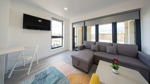 A modern living room with a sectional sofa, a flat-screen TV mounted on the wall, a small table, and a chair. - Ilford Apartments on the Lane (Ilford)