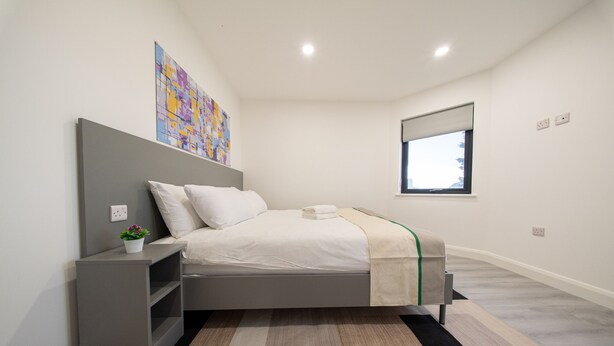 A modern bedroom with a bed, bedside table, and a window. - Ilford Apartments on the Lane (Ilford)