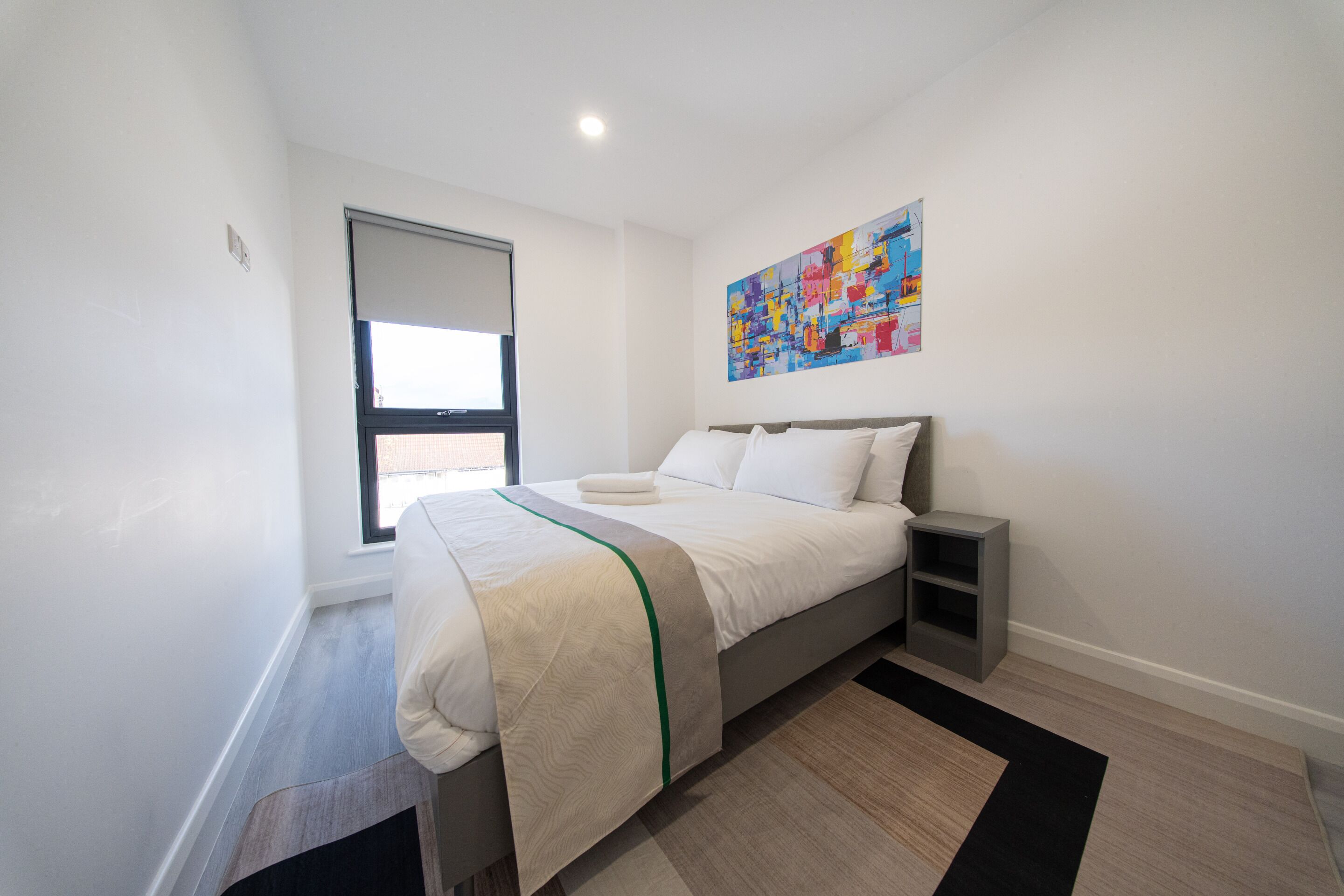 A modern bedroom with a bed, bedside tables, a window, and a colorful abstract painting on the wall.