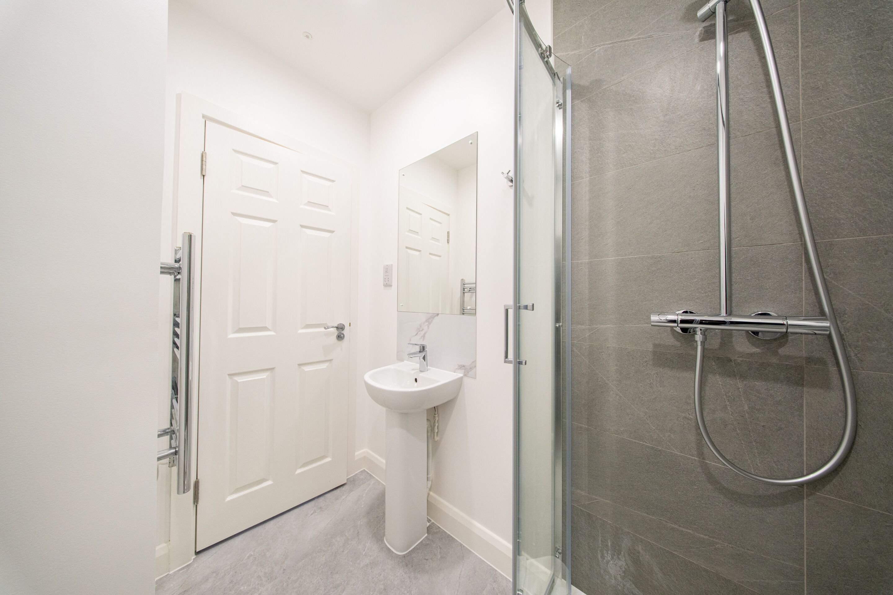 A modern bathroom with a glass-enclosed shower, a pedestal sink, and a white door.