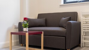 A grey sofa with a small wooden table in front, a potted plant, and a window with a view of trees. - Ilford Apartments on the Lane (Ilford)