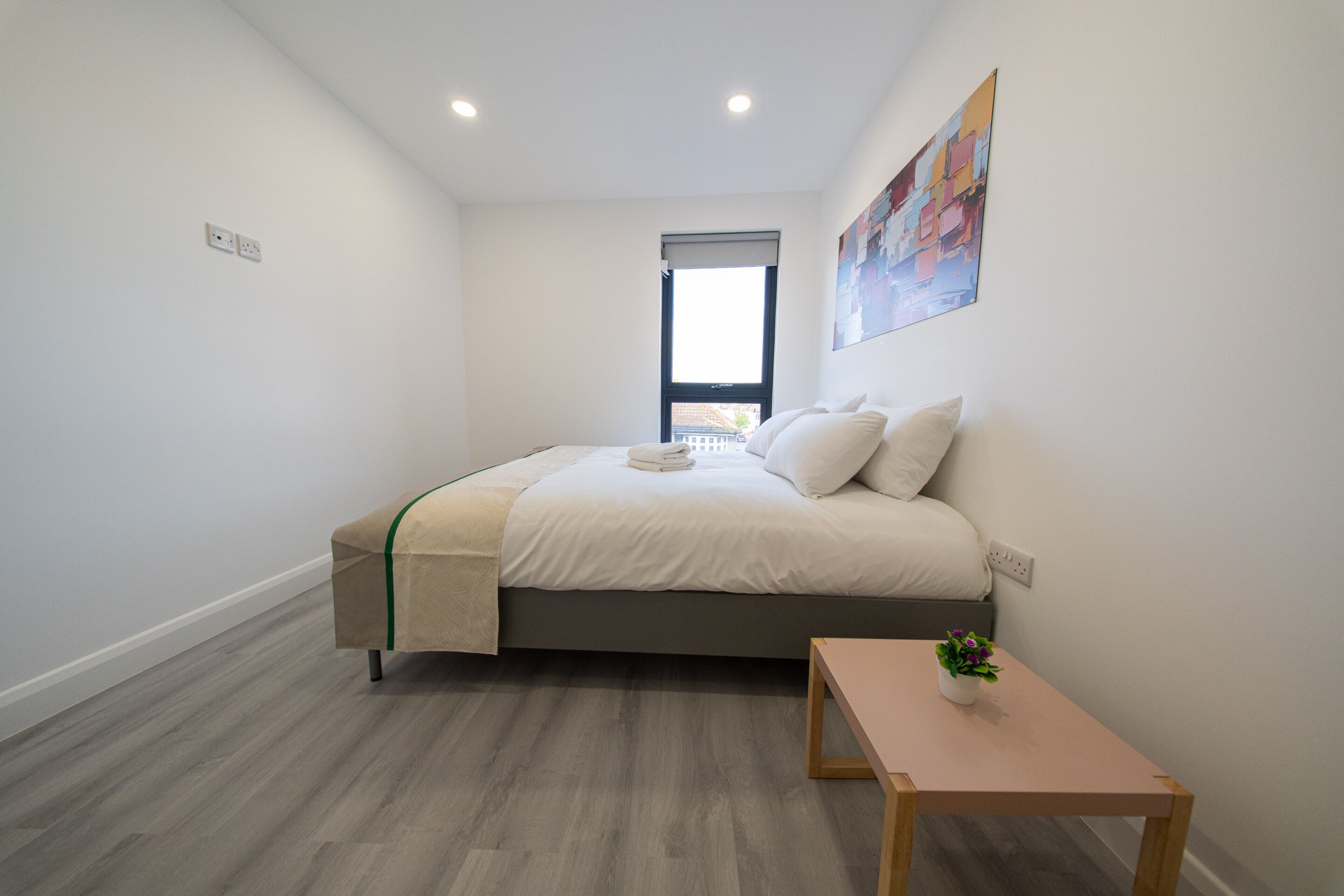 A bedroom with a bed, a small table, a window, and a painting on the wall.