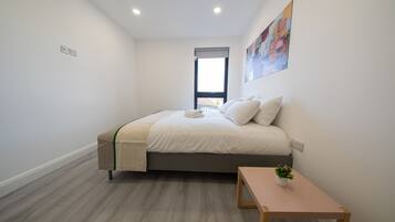 A bedroom with a bed, a small table, a window, and a painting on the wall.