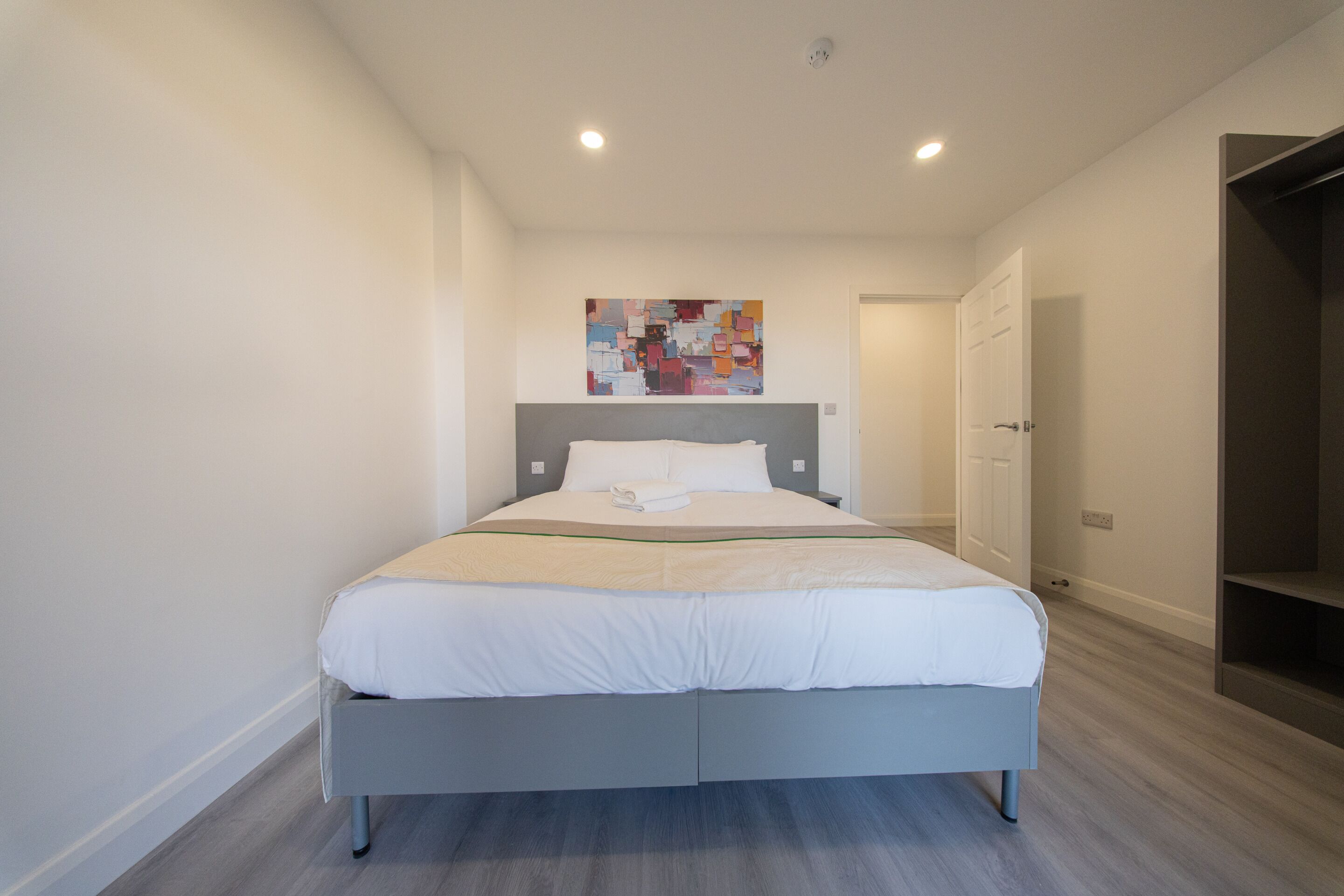 Un dormitorio moderno con una cama gris, ropa de cama blanca, un cuadro abstracto colorido en la pared y suelo de madera.