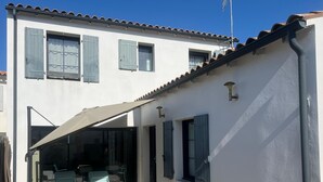 A covered outdoor patio with a table and chairs, a building with white walls and blue shutters, and a tiled roof. - Bright house in the heart of the village-100m from the ocean-6people (Angoulins)