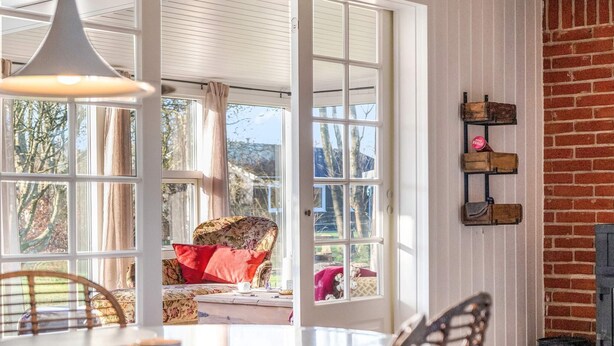 A room with a dining table, chairs, a hanging light fixture, a window with curtains, and a brick wall. - 4 Star Holiday Home in Hemmet (Hemmet)