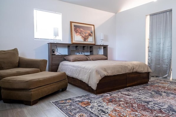 A bedroom with a bed, a sofa, a desk with a lamp, a framed picture on the wall, and a patterned rug.