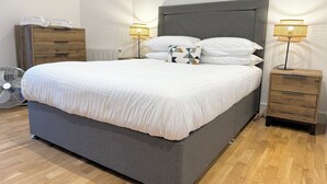 Una cama bien tendida con un cabecero gris, dos mesitas de noche con lámparas, un aparador de madera y un radiador.