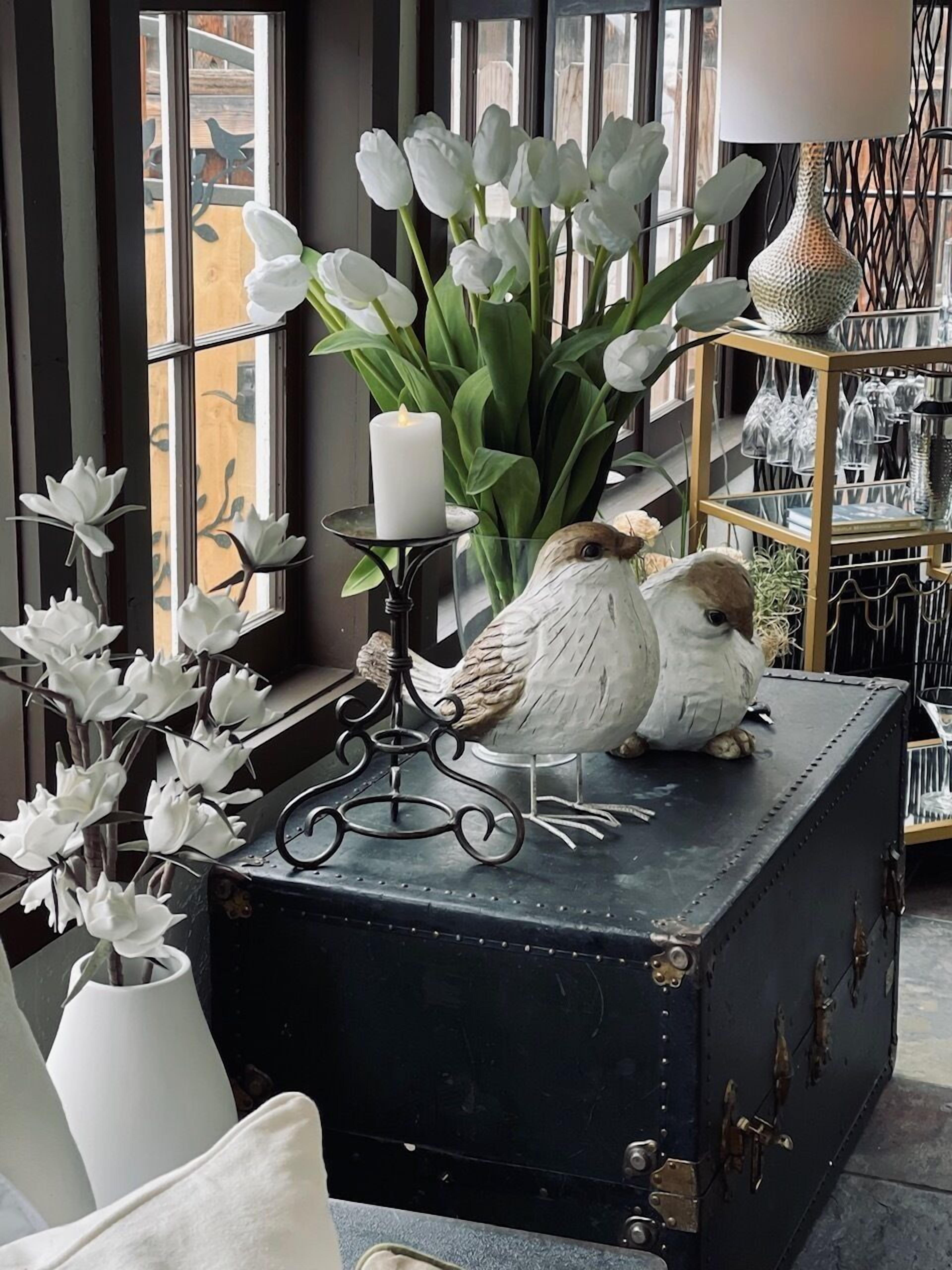 Een vintage koffer met decoratieve vogels en witte bloemen, geplaatst op een vensterbank met een kaars en een lamp.