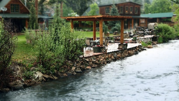 A riverside patio with outdoor seating and a gazebo, surrounded by lush greenery and wooden cabins. - The Cabins at Country Road (Evergreen)