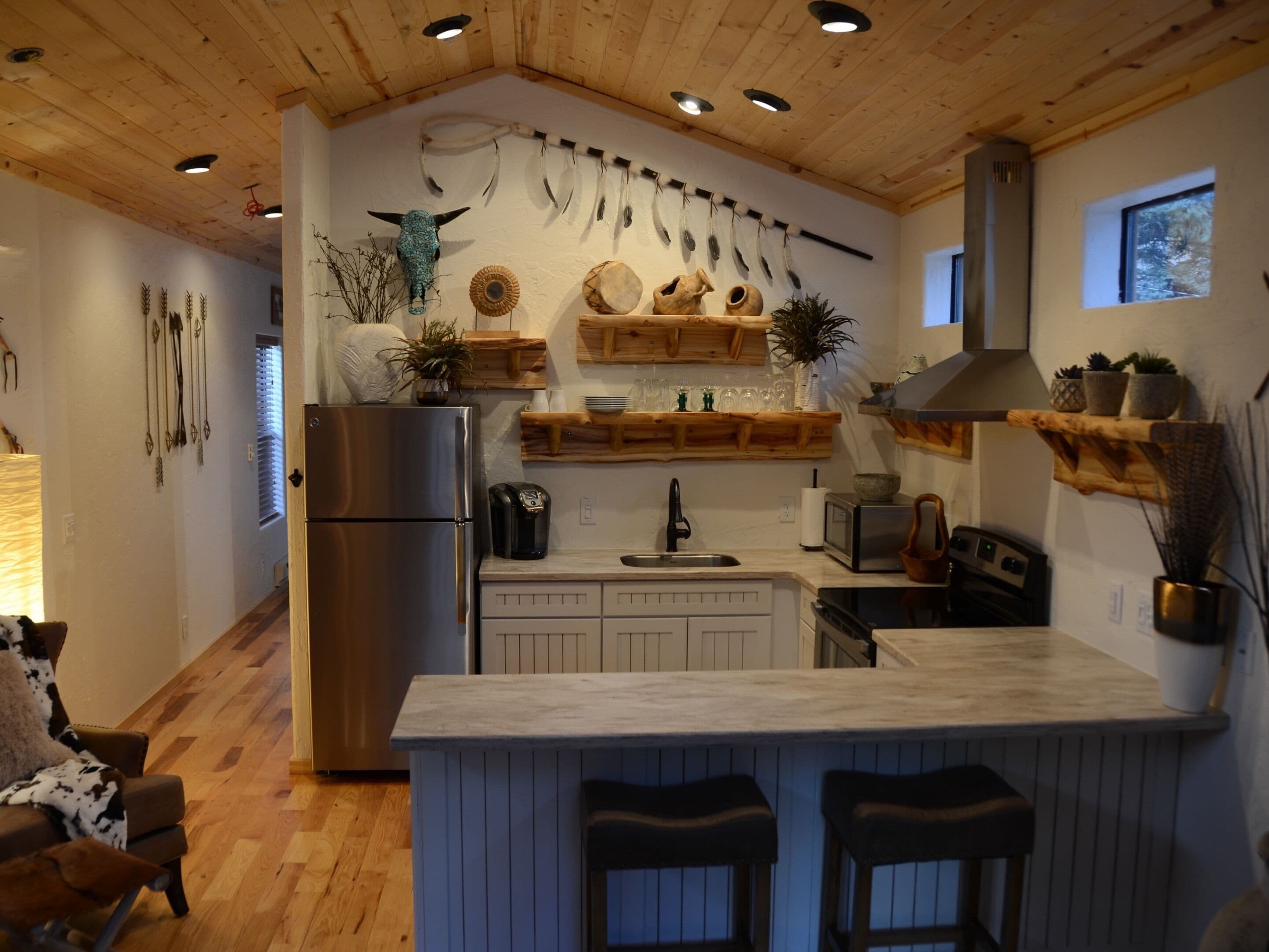 Une cuisine moderne avec des meubles en bois, un réfrigérateur en acier inoxydable, un îlot central intégré avec des tabourets de bar, et des éléments décoratifs tels que des plumes et des objets en céramique.