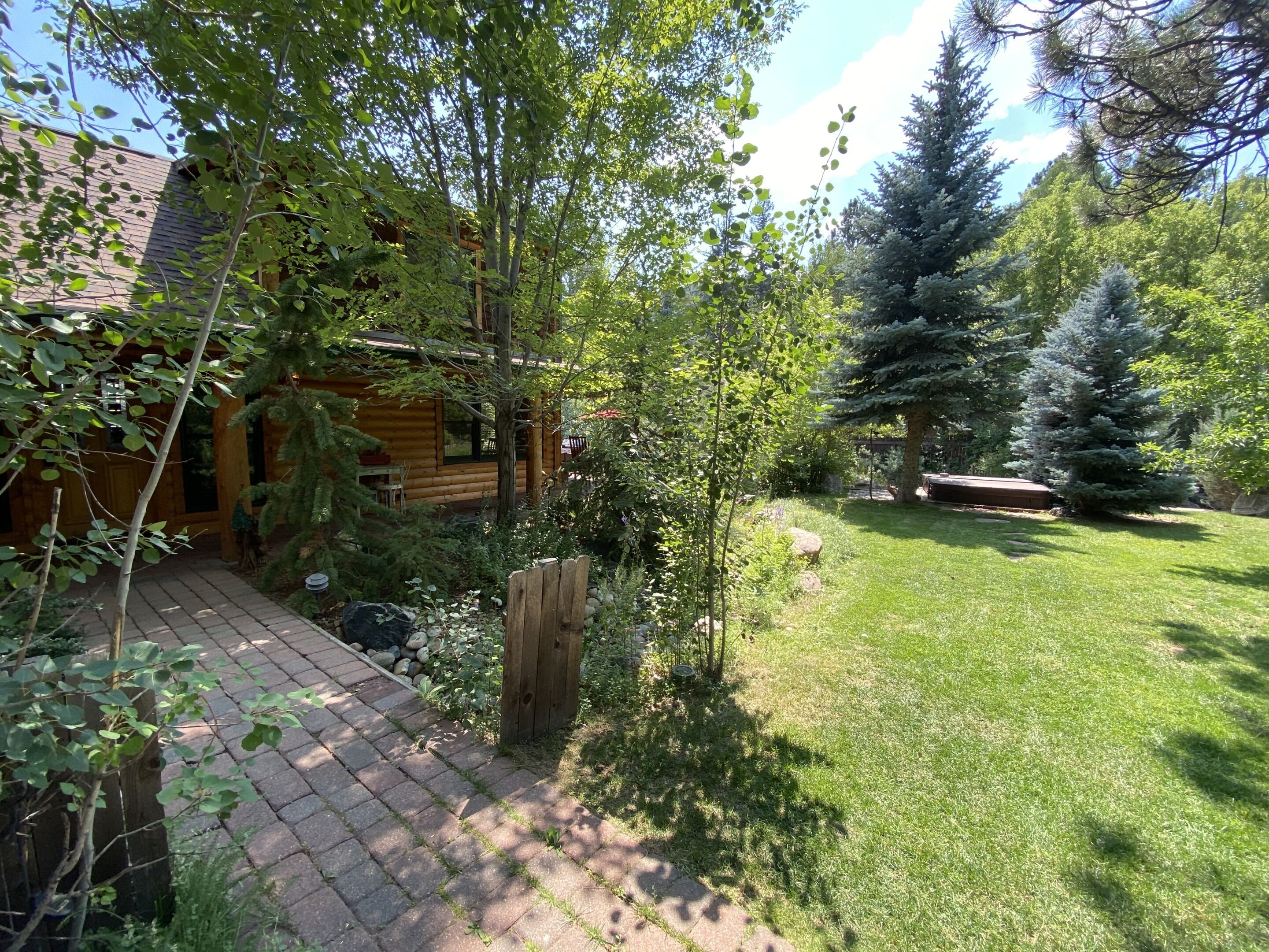 A wooden cabin with a brick pathway leading to the entrance, surrounded by lush greenery and a well-maintained lawn.