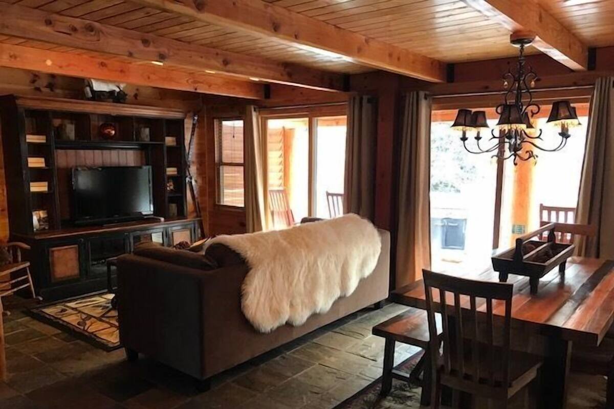 Interior acogedor de una cabaña con techo de madera, un televisor grande sobre un soporte de madera, un sofá con una manta de piel, una mesa de comedor con sillas y una araña.