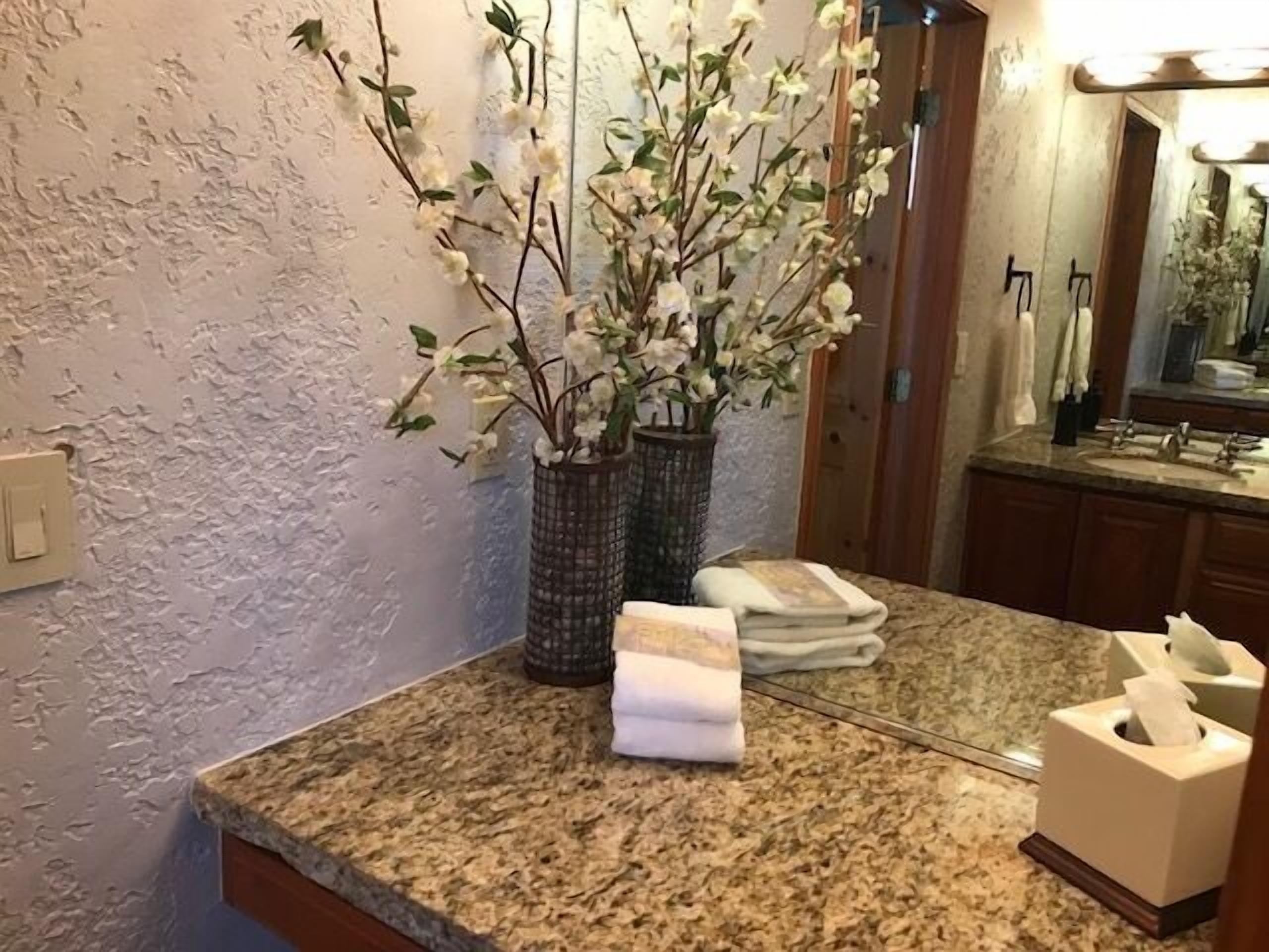 Ein Badezimmer mit einer Granit-Arbeitsplatte, zwei Vasen mit weißen Blumen, einem Handtuch und einer Taschentuchbox.