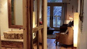 A bathroom with a large mirror, a sink, and a wooden vanity. There is a window with a view of trees outside. - The Cabins at Country Road (Evergreen)