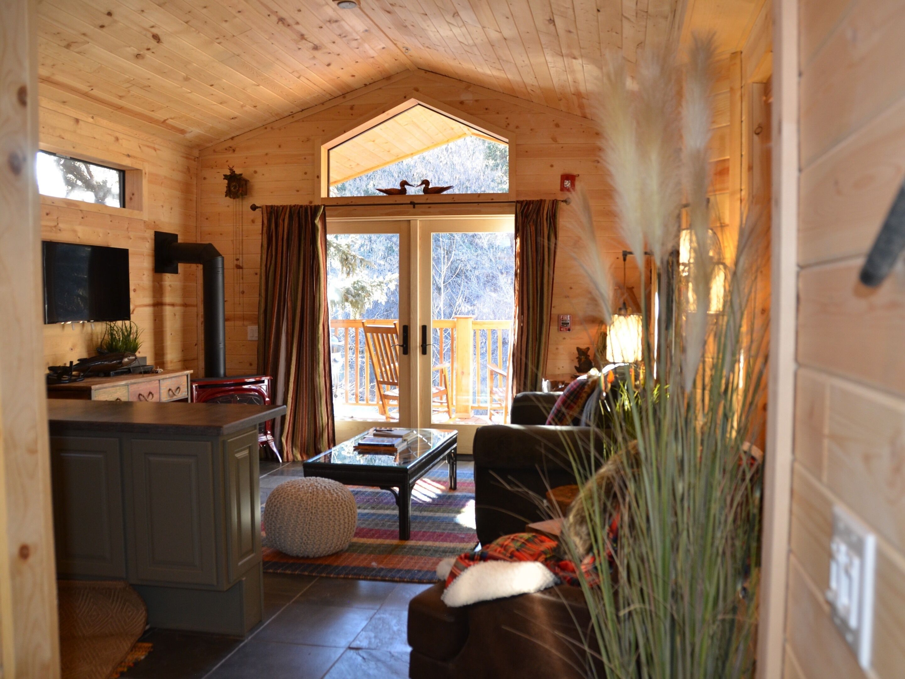A cozy cabin interior with a wooden ceiling, a fireplace, a dining area, and a large sliding glass door leading to a deck.