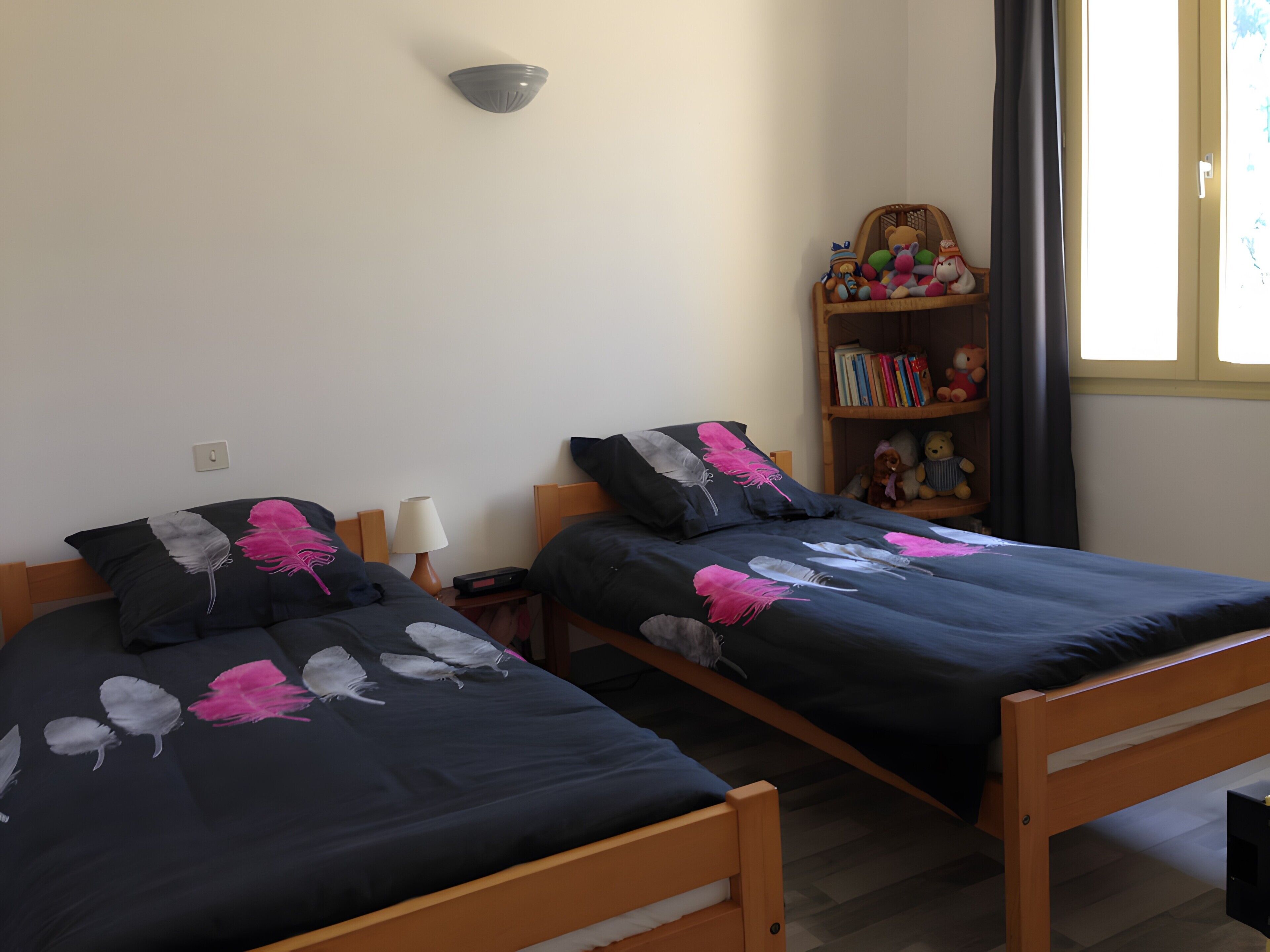 Zwei Einzelbetten mit federleichter Bettwäsche, ein Nachttisch mit Lampe, eine Wandleuchte und ein Bücherregal mit Stofftieren und Büchern.
