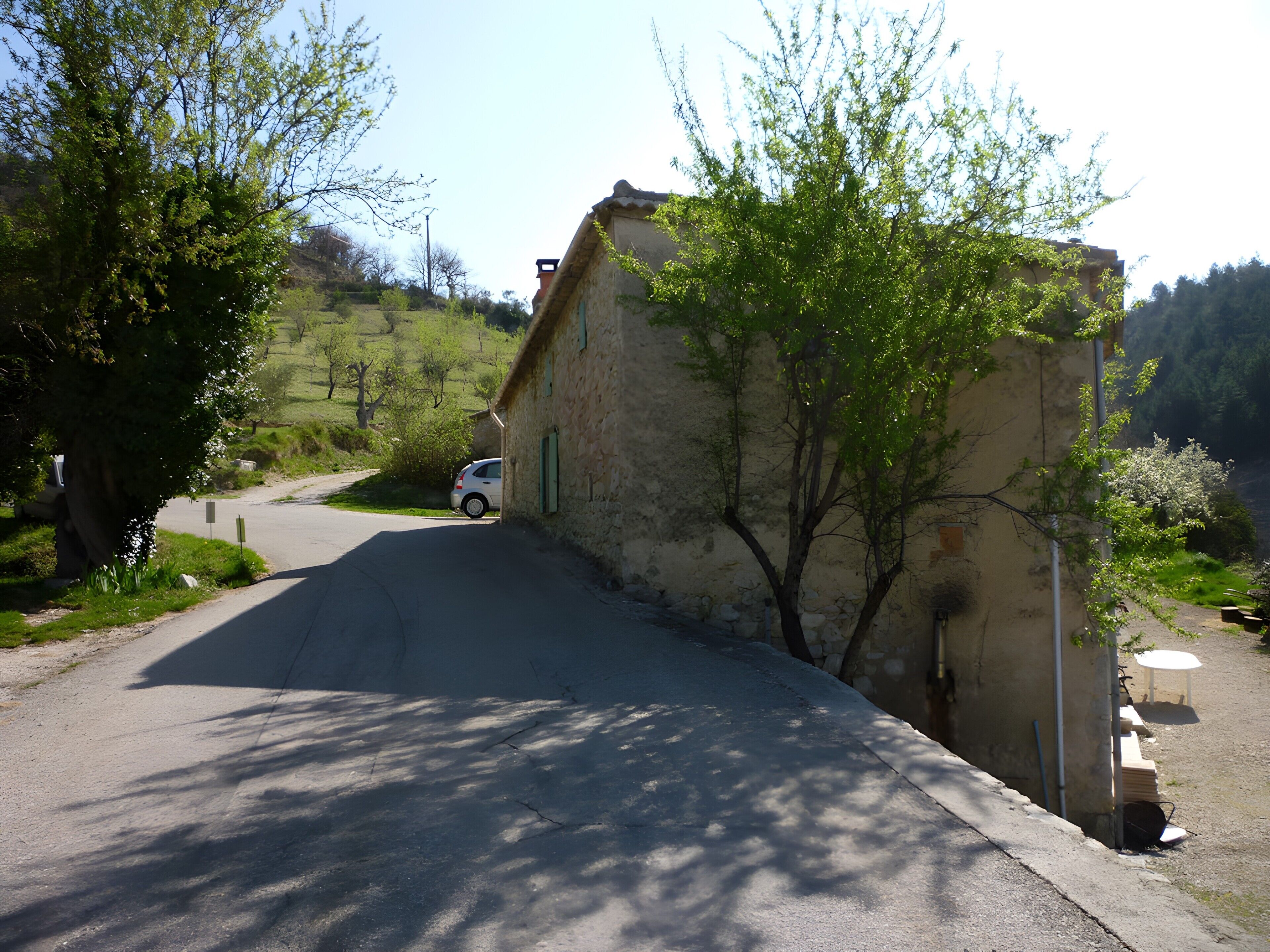 A narrow road with a parked car, a stone building, and trees.