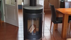 Un poêle à bois dans un salon, avec un sol carrelé, une table à manger et une cuisine en arrière-plan.