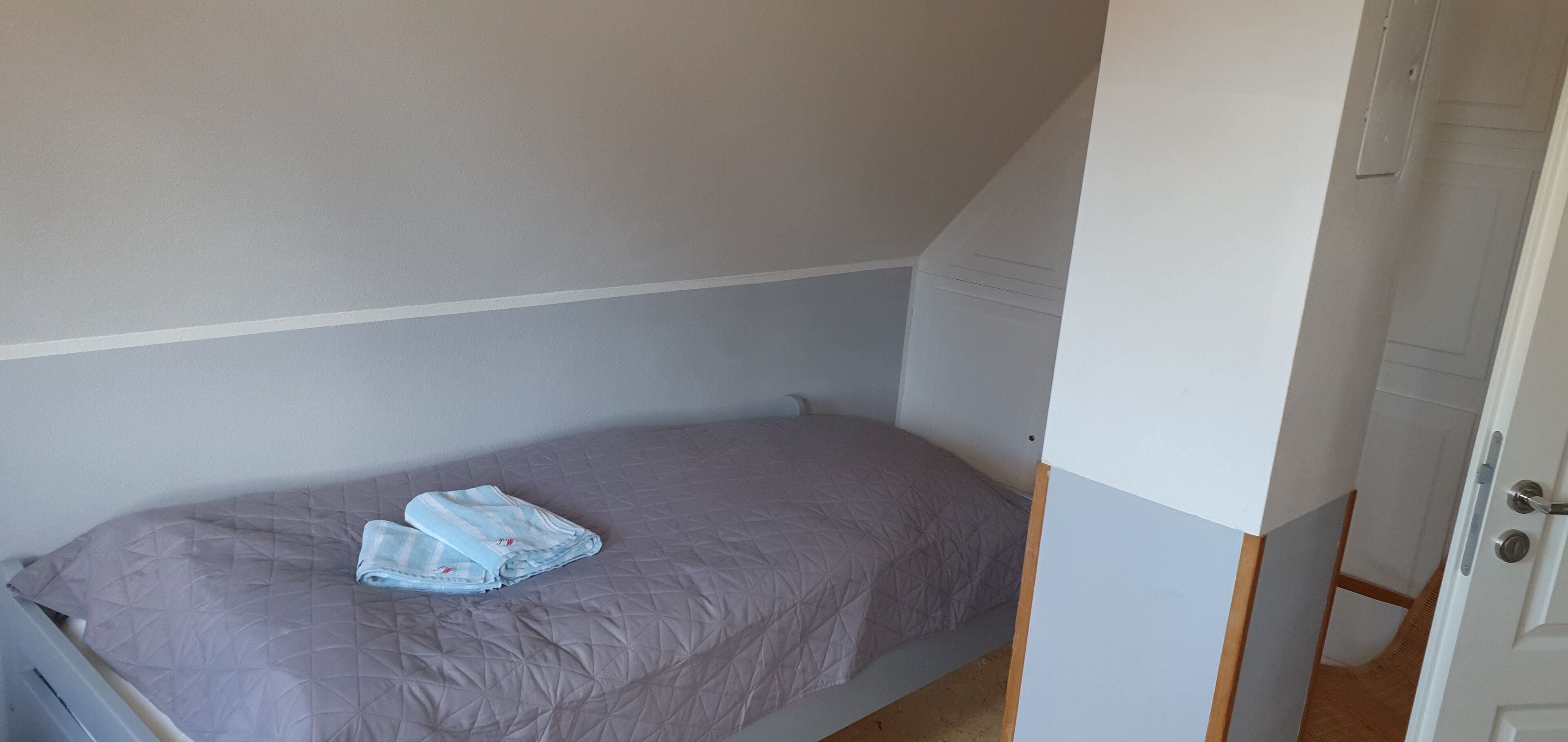 A small, single-bed room with a grey bedspread and a folded towel on the bed.