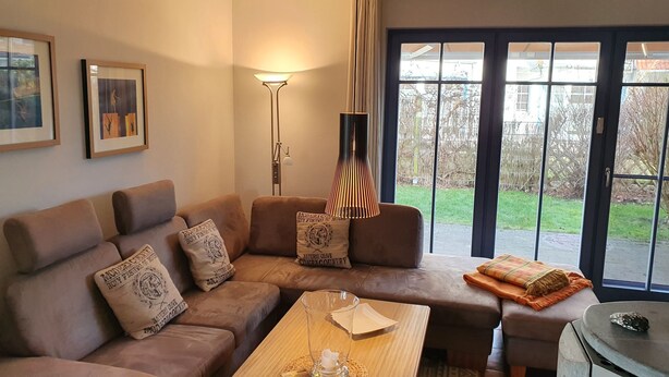 Un salon chaleureux avec un canapé d’angle marron, une table basse en verre et une vue sur un jardin, visible à travers de grandes portes vitrées.