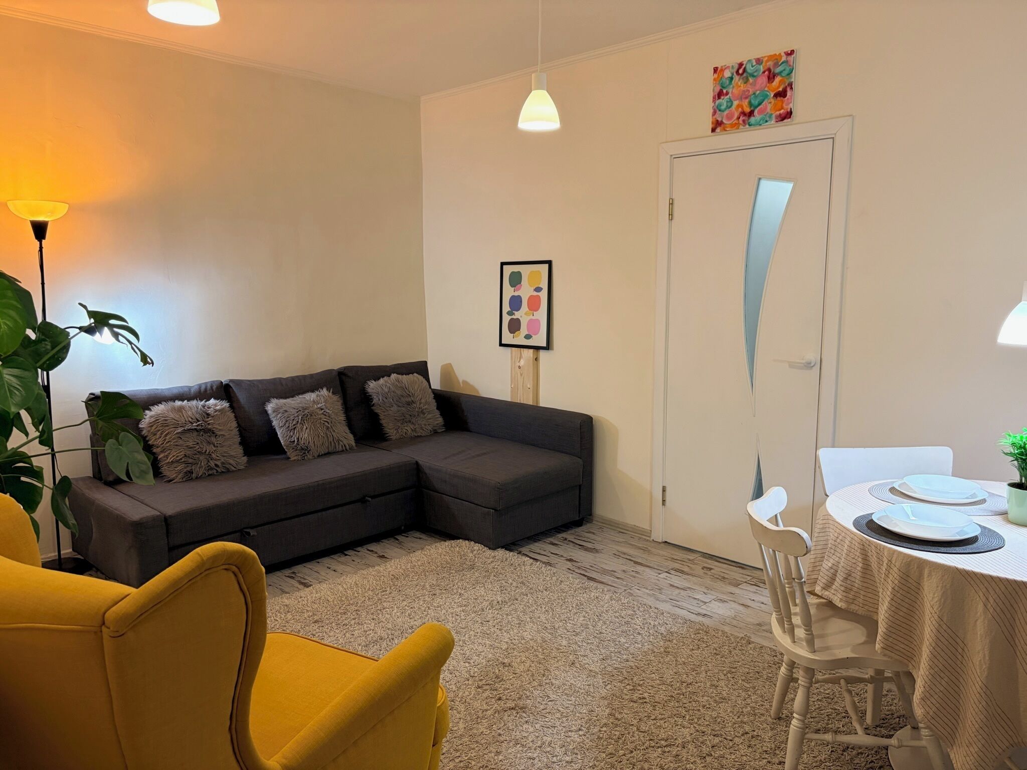 Ein Wohnzimmer mit einem grauen Ecksofa, einem gelben Sessel, einem Esstisch mit Geschirr und einer Pflanze, sowie einer Tür mit einem farbenfrohen Kunstwerk darüber.