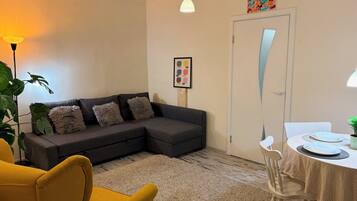Ein Wohnzimmer mit einem grauen Ecksofa, einem gelben Sessel, einem Esstisch mit Geschirr und einer Pflanze, sowie einer Tür mit einem farbenfrohen Kunstwerk darüber.