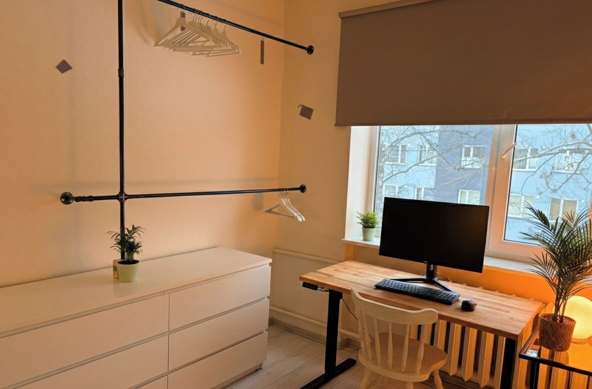 A small, well-lit room with a wooden desk, a computer, a chair, a radiator, a window with a view of buildings, and a plant on the desk.