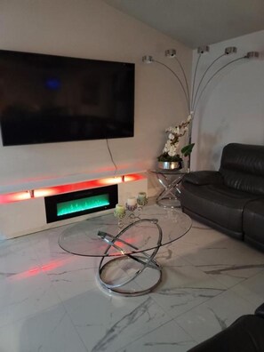 A modern living room with a flat-screen TV mounted on the wall, a glass coffee table with a ring light, a black leather sofa, and a small side table with a vase of flowers. - Stylish 3BR Pool Home Villa Frankely! (TAMPA)