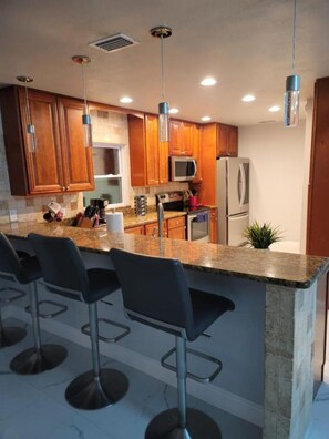 A modern kitchen with wooden cabinets, a granite countertop, stainless steel appliances, and bar stools. - Stylish 3BR Pool Home Villa Frankely! (TAMPA)