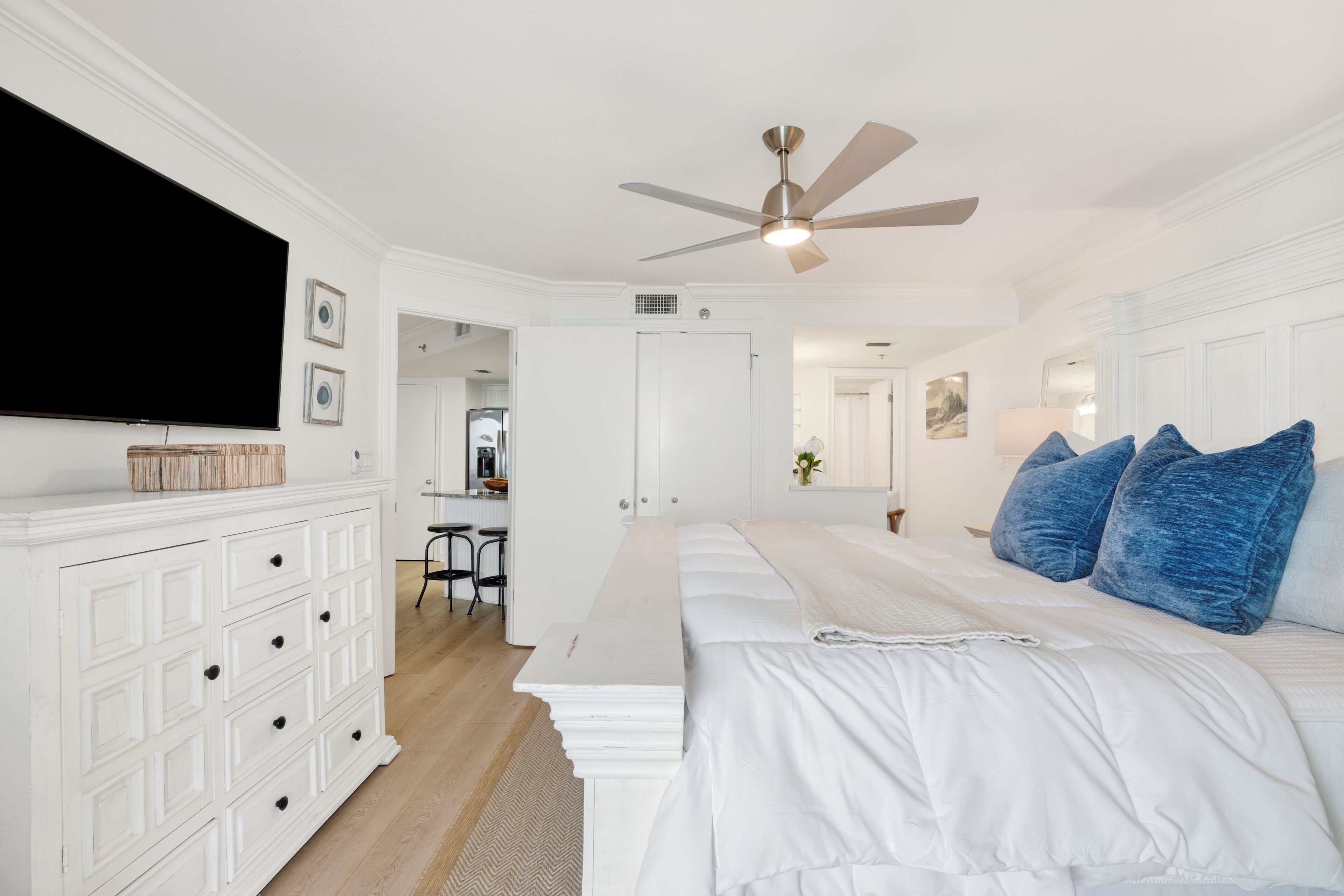 Ein Schlafzimmer mit einem Bett, einer weißen Kommode, einem Deckenventilator und einem Fernseher an der Wand.