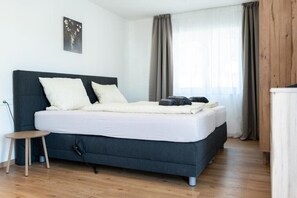A modern bedroom with a dark blue bed, white bedding, and a small wooden stool.