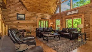 A log cabin interior with leather sofas, a coffee table, and large windows offering a view of greenery. - Luxury Cabin with Private Waterfall & Hot Tub (Sevierville)