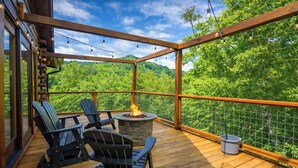 A wooden deck with Adirondack chairs, a stone fire pit, and a view of a forested hillside. - Luxury Cabin with Private Waterfall & Hot Tub (Sevierville)