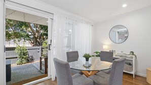 Un área de comedor con una mesa de vidrio y cuatro sillas grises, una pared blanca con un espejo redondo, y vistas a un árbol y una valla a través de un vidrio.