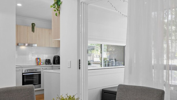 Una cocina moderna con armarios blancos, horno de acero inoxidable y cafetera. Hay un comedor con una mesa de vidrio y dos sillas.