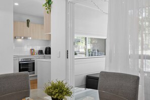 A modern kitchen with white cabinets, a stainless steel oven, and a coffee maker. There is a dining area with a glass table and two chairs. - Bright 3-bed Home Near Maroochydore Beach (Sunshine Coast)