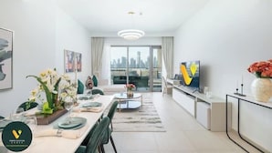 A modern living room with a dining area, a flat-screen TV, and a city view through large windows. - Vogue | Downtown with Direct Access to Dubai Mall (Dubai)