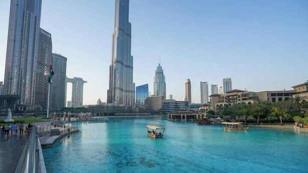 A waterfront cityscape with modern skyscrapers, including a prominent tall building, and a calm river with boats. - Vogue | Downtown with Direct Access to Dubai Mall (Dubai)