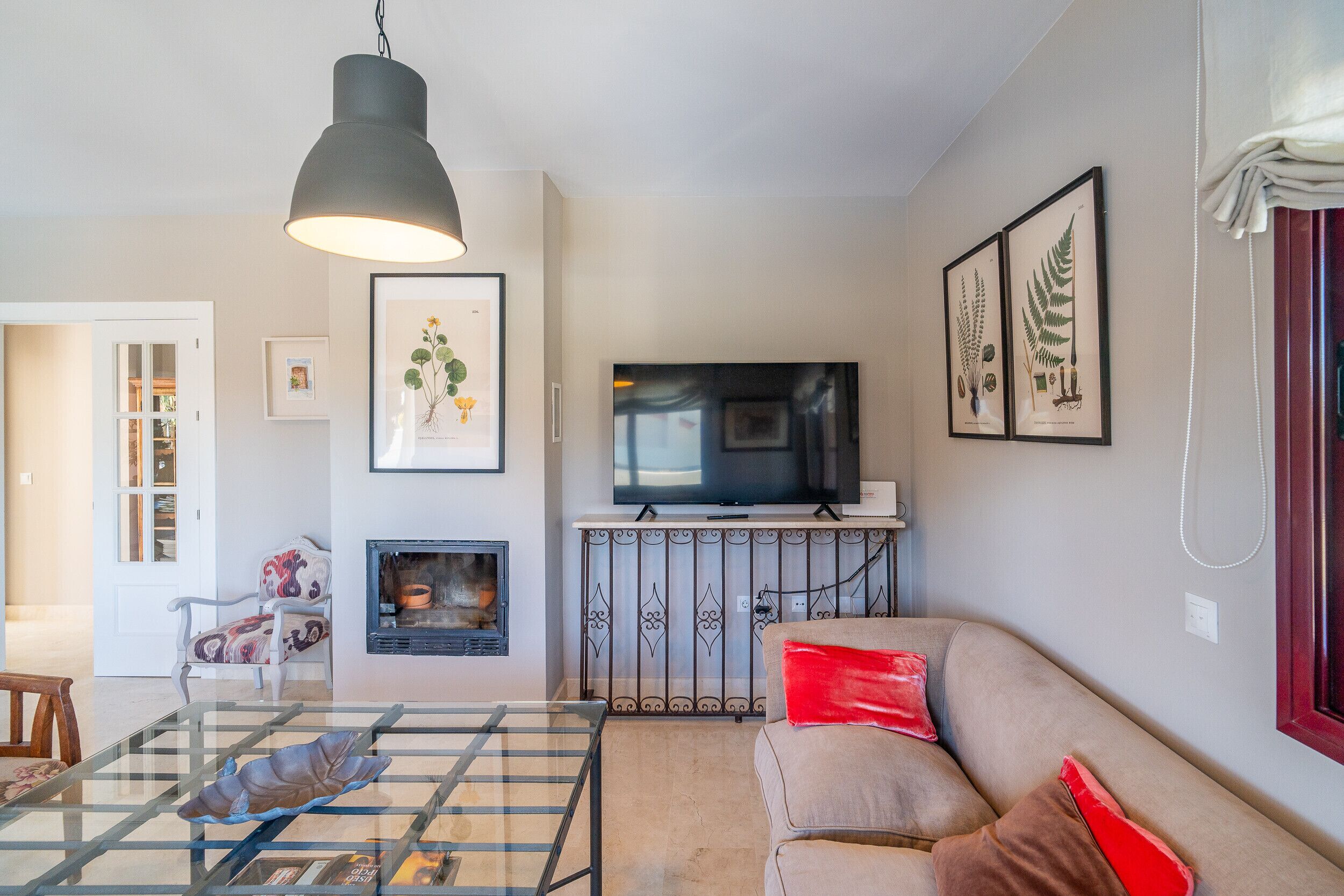 A living room with a sofa, a glass coffee table, a fireplace, a television, and framed botanical prints on the wall.