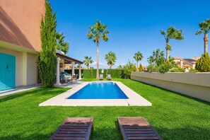 Una piscina con due lettini in primo piano, circondata da erba curata e palme.